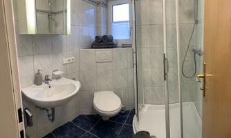 A modern bathroom with a shower, a sink, and a toilet. The walls are tiled in light colors, and there are fresh towels on the shelf.