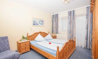 A cozy bedroom with a wooden double bed and blue bedding. The room has large windows with curtains and is bright and cheerfully furnished.