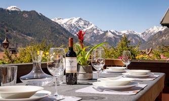 A beautiful set table on a terrace overlooking snow-covered mountains. A bottle of wine and glass decorations enhance the inviting atmosphere.