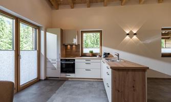 A modern kitchen with bright furniture and a wooden ceiling. Large windows provide plenty of daylight and a view of nature.