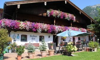 A traditional farmhouse with colorful flowers in boxes and a well-kept garden. In the foreground, there are seating furniture and a sun umbrella.