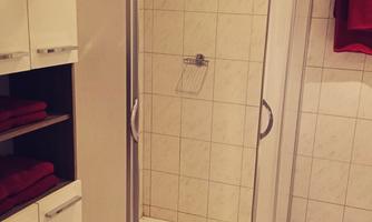 A modern shower with a glass door in a bright bathroom. Tiled walls all around and a shelf with towels complement the space.