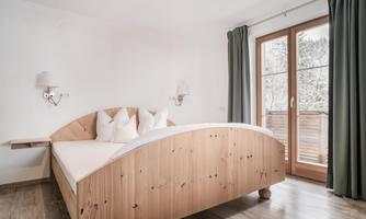 A bright bedroom with a wooden bed and white pillows. Large windows with curtains provide a view outside.