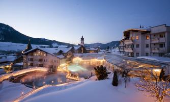 A picturesque winter village with snow-covered roofs and gentle hills. The outdoor baths are bathed in soft light while the evening sky is twilight.