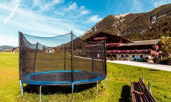 A trampoline is standing in a green meadow with mountains in the background. It is a sunny day with a blue sky and a few clouds.