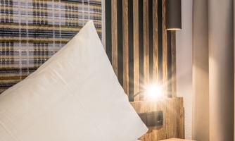 A cozy bedroom with a large beige pillow and modern wood details. Next to the bed there is a gentle light.