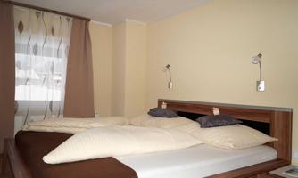 A modern bedroom with a double bed and beige walls. There are windows with curtains and wall sconces next to the bed.