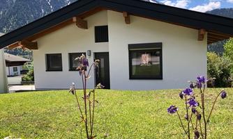 A modern house with a garden and colorful flowers. In the background, the mountains and a clear sky can be seen.
