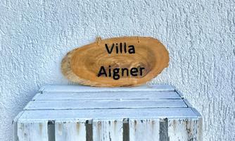 A wooden sign with the inscription "Villa Aigner" hangs on a white wall. It stands on a white wooden table.