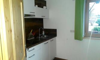 A small, modern kitchen with white cabinets and a wooden surface. The room has a window with green curtains and offers plenty of daylight.