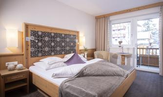 A cozy bedroom with a large bed and wooden decor. The room features a balcony with a view and warm colors.