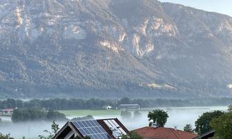 A sunny valley with mountains in the background and a calm area. In the foreground, houses with solar panels are visible.