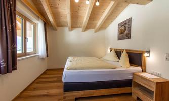 A cozy bedroom with a double bed and wooden floors. The walls are light and there are large windows that provide plenty of light.