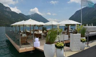 A stylish restaurant by the water with sun umbrellas and modern furniture. In the background, mountains and a clear sky can be seen.