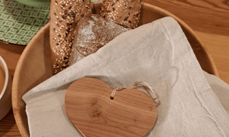 A wooden board with freshly baked bread and a decorative wooden heart. The bread is partially sprinkled with sesame seeds and is resting on a white cloth.