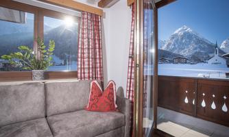 A cozy apartment with a gray sofa and red cushions. The view from the window shows snow-covered mountains and a clear sky.