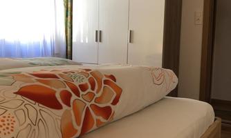 A bright bedroom with a bed and a colorful duvet. In the background, there is a wardrobe and a window with curtains.
