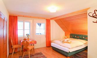 A cozy room with a double bed and a small dining table. It has large windows and warm wooden details.