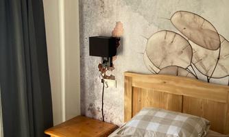A cozy bedroom with a wooden bed and decorative cushions. The wall is adorned with a soft, artistic design and there is a nightstand with a lamp.