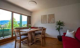 A bright and modern living room with a wooden dining area. Large windows offer a view of nature and plenty of daylight.
