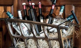 A wooden cart is filled with various bottles, including wine and spirits. The background is made of wood, which gives the image a rustic atmosphere.