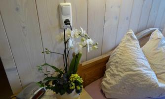 A pretty flower arrangement with white orchids and green plants sits on a nightstand. The background features light wooden panels and a cozy pillow.