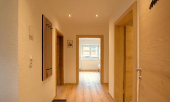 A bright hallway with a wooden floor and white walls. Several doors lead to adjacent rooms and there is a window view at the end of the hallway.