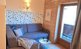 A cozy living room with a gray sofa and wooden details. The room has access to the balcony and a decorative wall with a plant pattern.