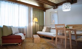 A cozy living space with a wooden ceiling and a large window. The room is furnished with a sofa, a dining table, and a lamp that creates an inviting atmosphere.