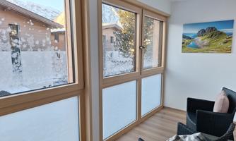 A cozy room with large windows that offer a view of snow-covered landscapes. On the windowsill sits a picture of mountains and green meadows.