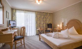 A cozy hotel room with a double bed, a desk, and wooden furniture. Large windows let in plenty of light and offer a view of the floral curtains.