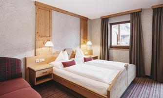 A cozy bedroom with two single beds and wooden decor. The room has a window, a comfortable chair, and warm colors.