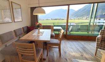 A modern living space with a wooden table and chairs. Through the large window, one looks out onto a picturesque mountain landscape.