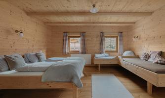 A cozy wooden bedroom with two beds and bright windows. The walls and ceiling are made of wood, creating a warm atmosphere.