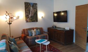A cozy living room with a sofa and a television on the wall. A large picture of a landscape and a wooden table complete the decor.