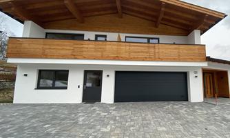 A modern house with a wooden roof and a large balcony. The driveway is paved with stone.