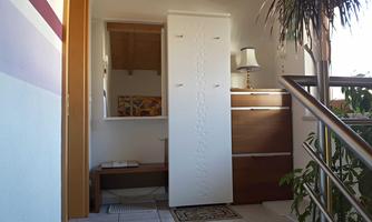 A bright hallway with a white cabinet and a wooden shelf. In the background, plants and a lamp are visible.
