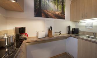A modern kitchen with wooden cabinets and a picture of a sunlit forest on the wall. On the countertop, there is a coffee machine and additional kitchen appliances.