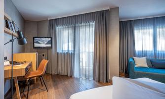 A modern hotel room with a large window and shimmering curtains. There is a desk, a chair, and a cozy couch in the room.