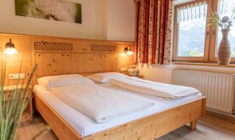 A cozy bedroom with a wooden bed and two mattresses. The room has bright walls and large windows with a view of the mountains.