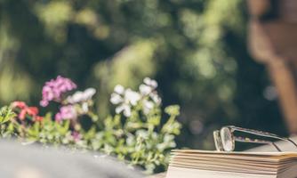 An open book lies on a table, next to a pair of glasses and colorful flowers. The scene feels calm and inviting, perfect for reading outdoors.