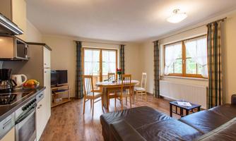 A cozy living room with a kitchenette, a dining table, and a sofa. Large windows let in a lot of light.