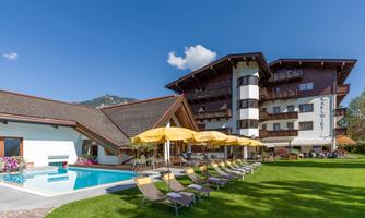 A beautiful hotel with an outdoor pool and sun loungers in the garden. The surroundings are picturesque with mountains and blue skies.