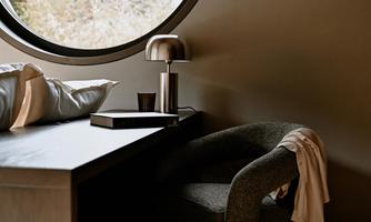 A cozy room with a round window that offers a view of nature. A stylish chair and a table with a lamp and small items give the room a modern atmosphere.