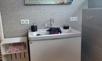 A modern bathroom with a white sink and a large mirror. It is bright and well-lit, with a wooden shelf on the left side.