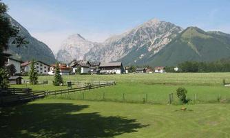 A picturesque landscape with green meadows and mountains in the background. In the foreground, some houses and trees are visible.