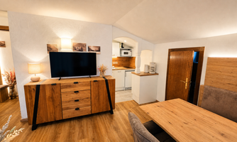 A modernly furnished living space with wooden surfaces and a large television. The kitchen is bright and well-equipped.