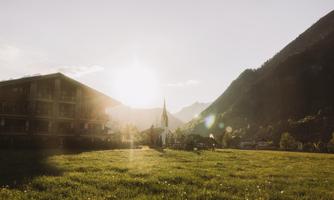 A picturesque landscape with gentle hills and a church in the foreground. The sun shines behind the mountains, bathing the scene in warm light.