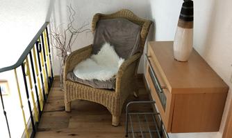 A cozy room with a rattan armchair and a wooden shelf. On the wall is a vase with a plant and a small decorative element.