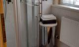 A modern bathroom with a triangular shower cabin and a trash can. Light streams through a window, creating a bright atmosphere.